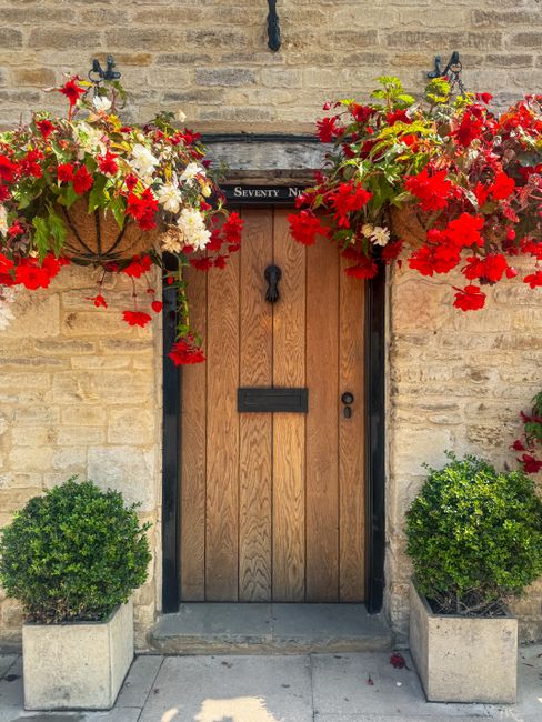 A rustic wooden door with a black metal handle and letterbox, set in a beige brick wall. Above the door, there are two large hanging baskets filled with vibrant red and white flowering plants, likely 
