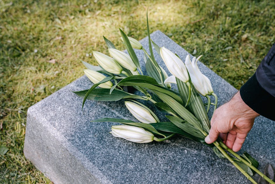 A hand is placing a bouquet of white lilies with long green leaves on a grey granite memorial stone, set outdoors on a grass surface. The lilies are fresh with smooth petals and sturdy stems, arranged