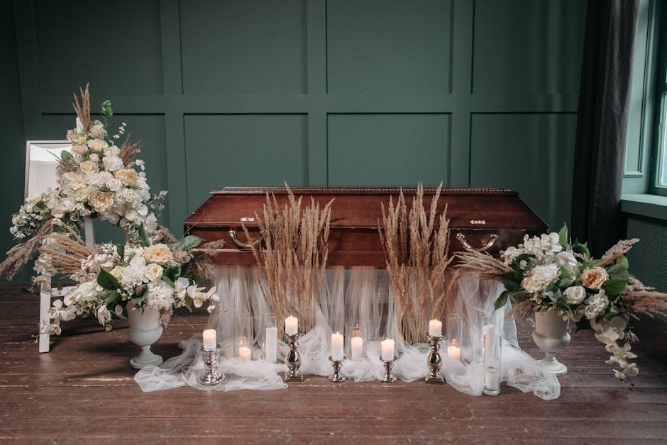 A floral arrangement placed on top of a vintage wooden casket during a funeral service, featuring cream and white roses, hydrangeas, and lilies, complemented by beige pampas grass and greenery. The fl