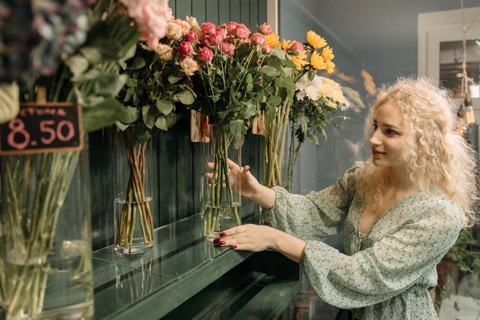 A woman with curly blonde hair and a light green, long-sleeved, floral dress is selecting freshly cut flowers from glass vases displayed on a dark green shelf at a florist shop. The vases contain a va